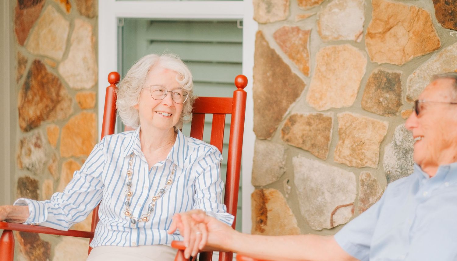 Elderly couple holding hands and laughing together on a porch with red rocking chairs and stone wall.