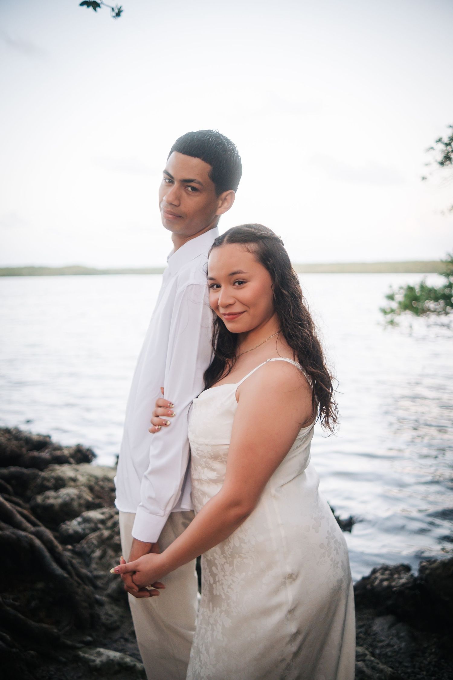 A couple in wedding attire embraces on a rocky shoreline at sunset with ocean waves in the background.