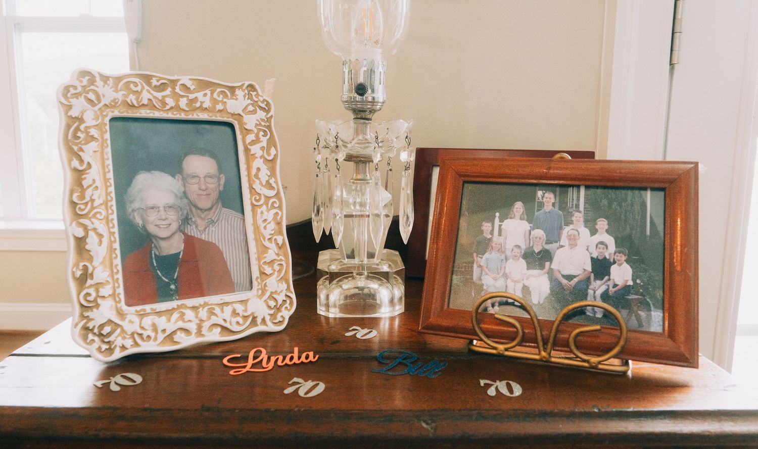 Two framed family photos displayed on a wooden table with a crystal lamp and a decorative Linda sign celebrating a 70th birthday.