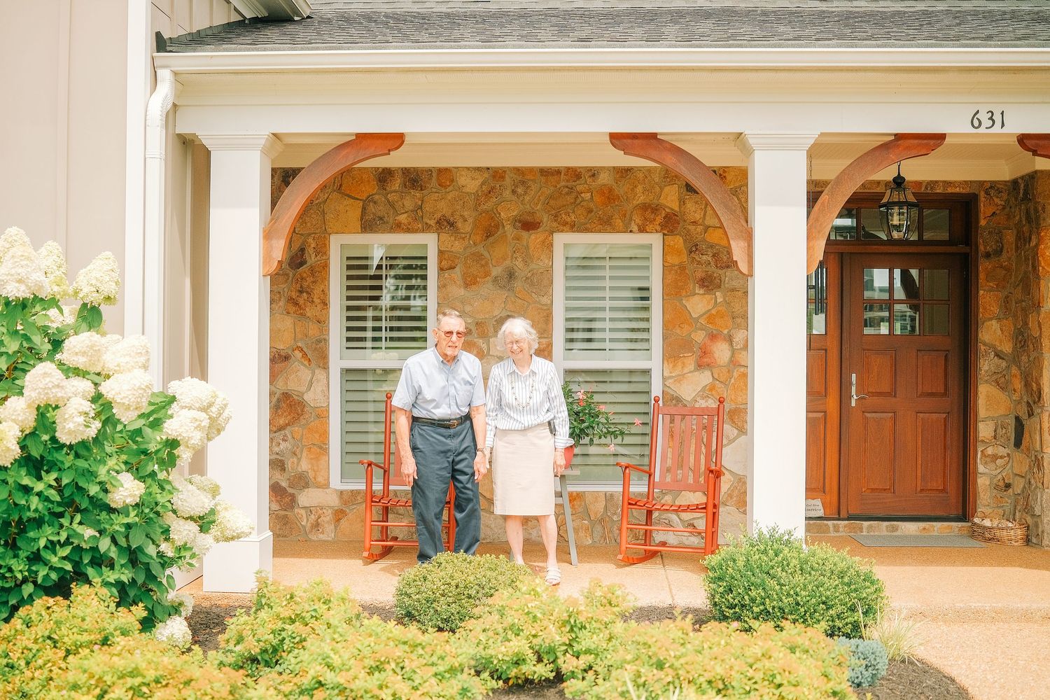 Elderly couple standing on stone porch of home numbered 631, surrounded by lush greenery and red rocking chairs.