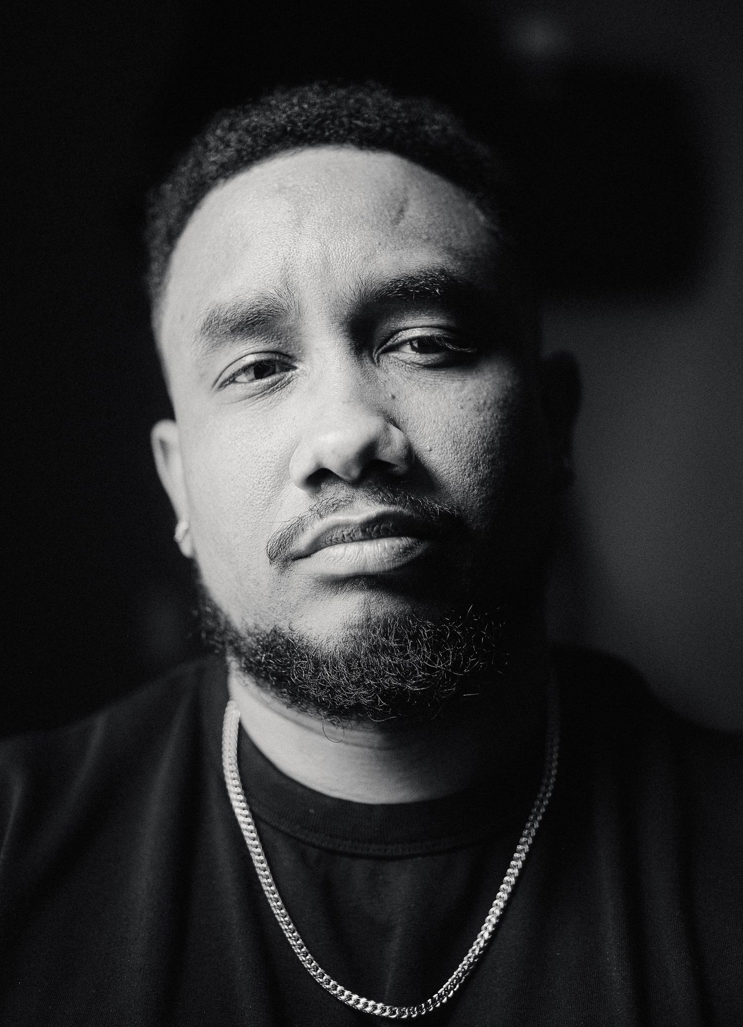 Black and white portrait of a young man with a beard wearing a chain necklace and black shirt.