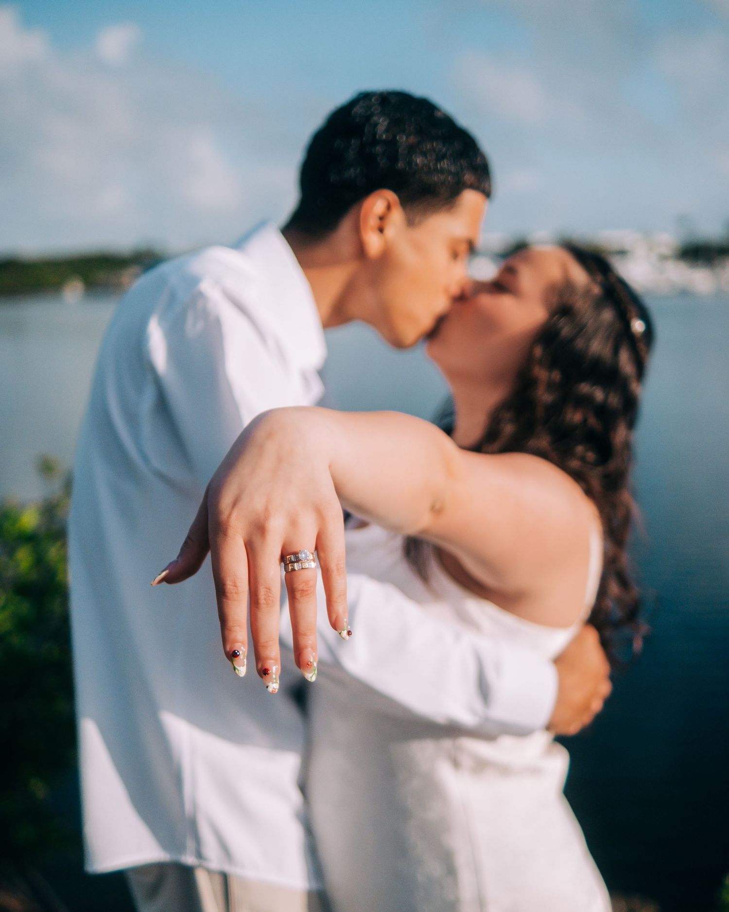 A couple embraces and kisses by the water in a romantic photoshoot showing off an engagement ring.
