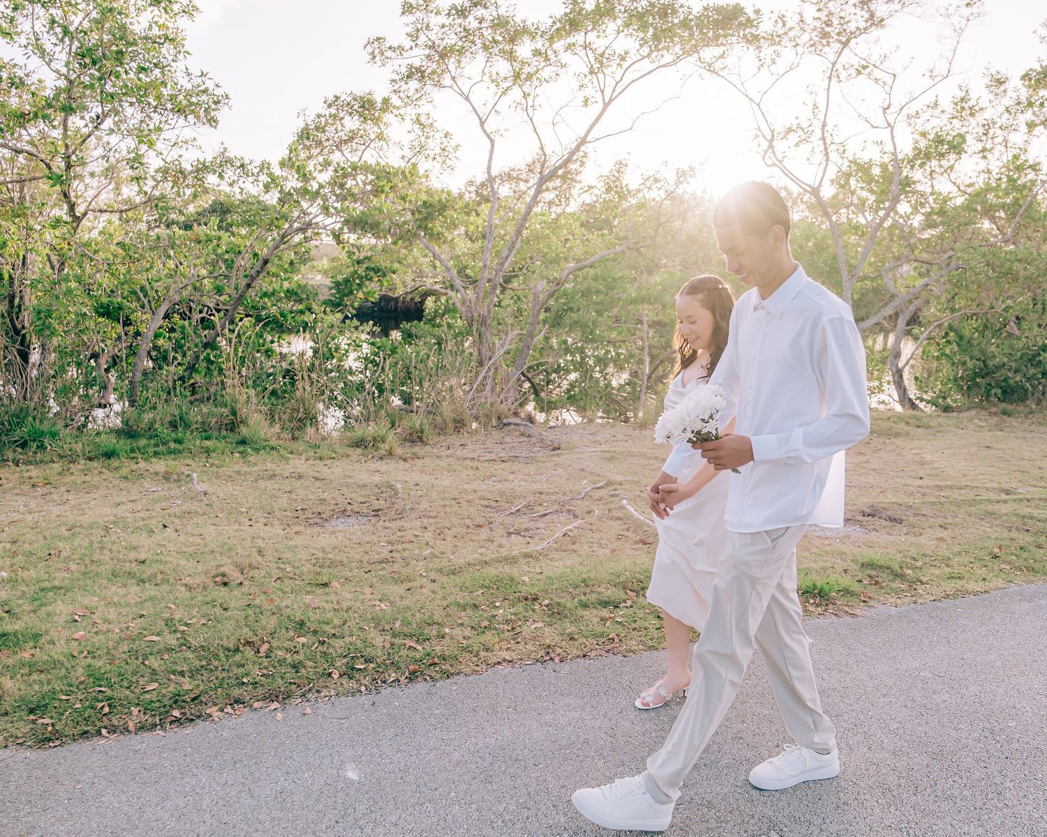 Romantic couple in white outfits walking along a sunlit path through nature during golden hour.