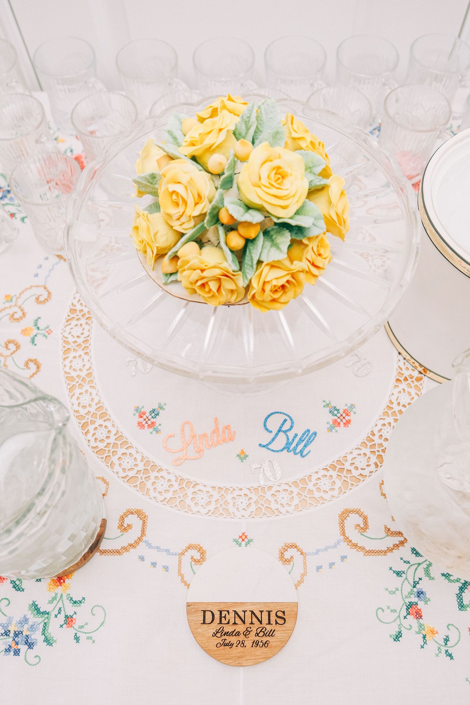 Elegant wedding table with yellow rose cake, embroidered linen, and wooden name card reading Dennis, Linda & Bill.