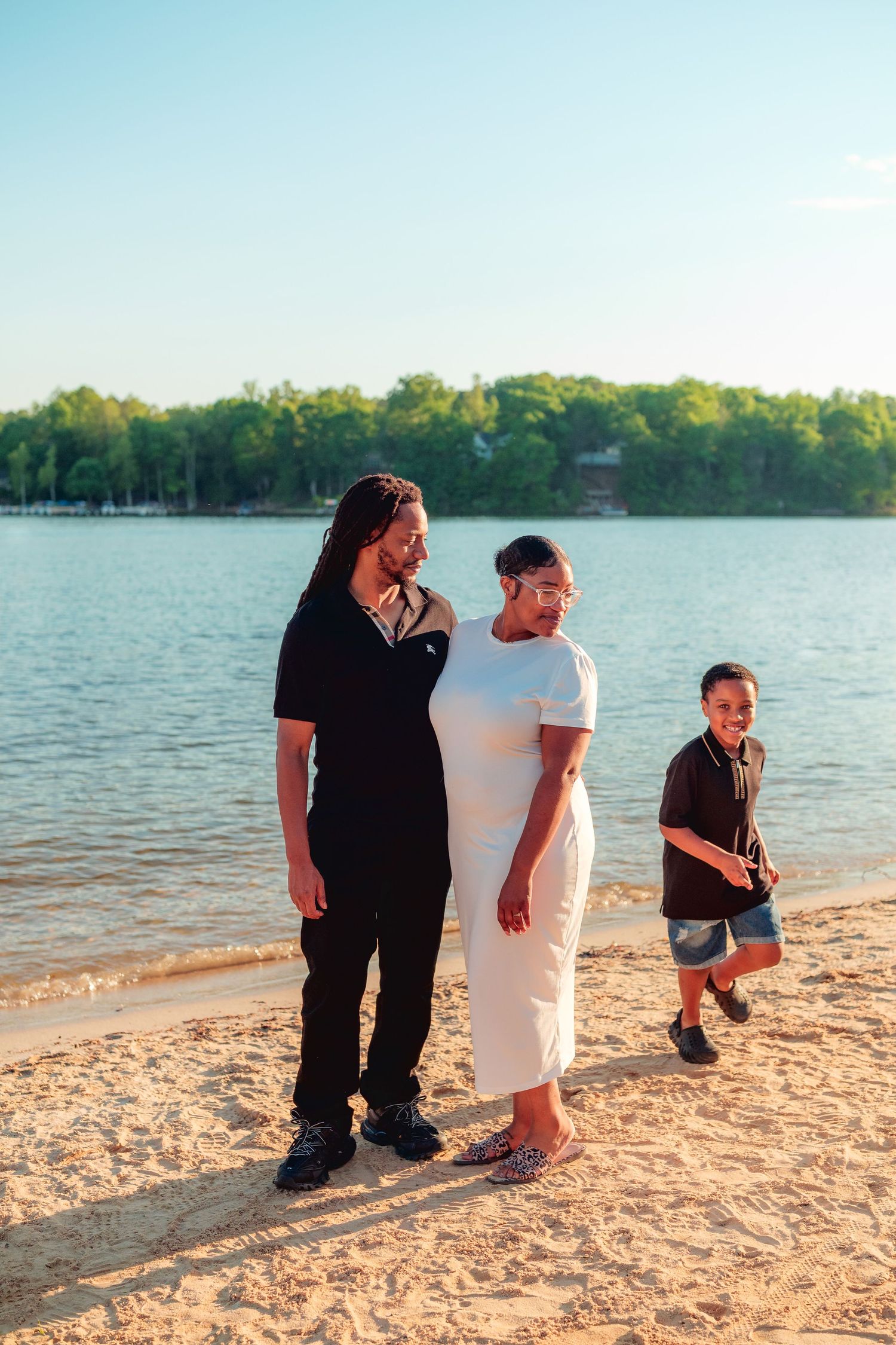 Family of three enjoying a sunny day on a sandy beach by a lake, with lush green trees in the background.