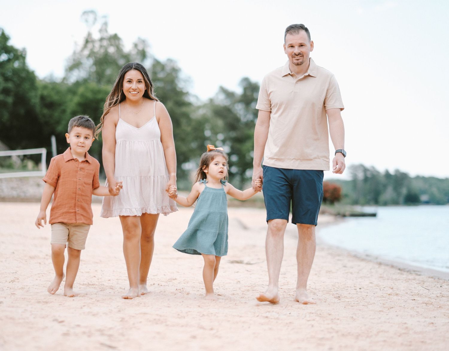 Family of four walking hand-in-hand on a sandy beach, smiling and enjoying a sunny day together.