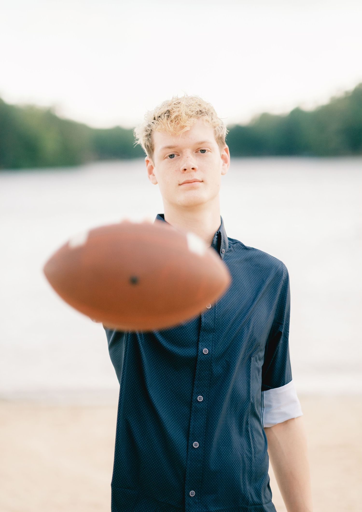 Young blonde teen holding a football toward camera near a lake, wearing a navy blue shirt.