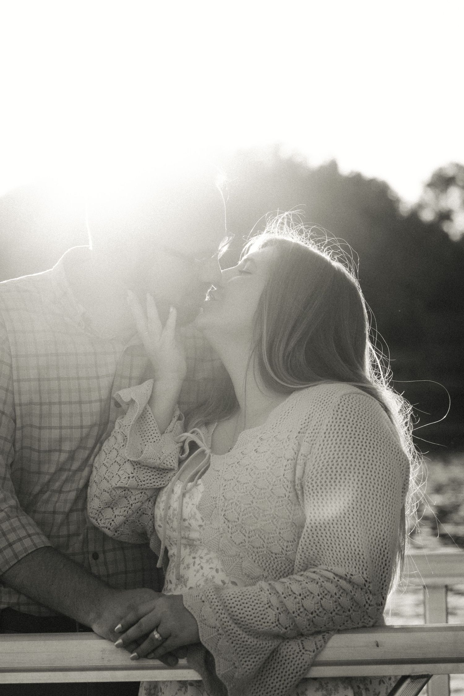 Black and white photo of a couple sharing a tender kiss in bright sunlight on a wooden bridge.
