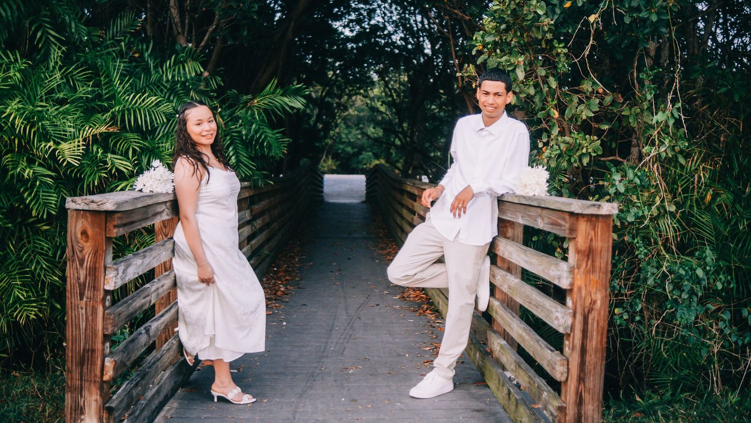 A couple in white outfits poses together in a lush green garden setting for romantic outdoor photos.