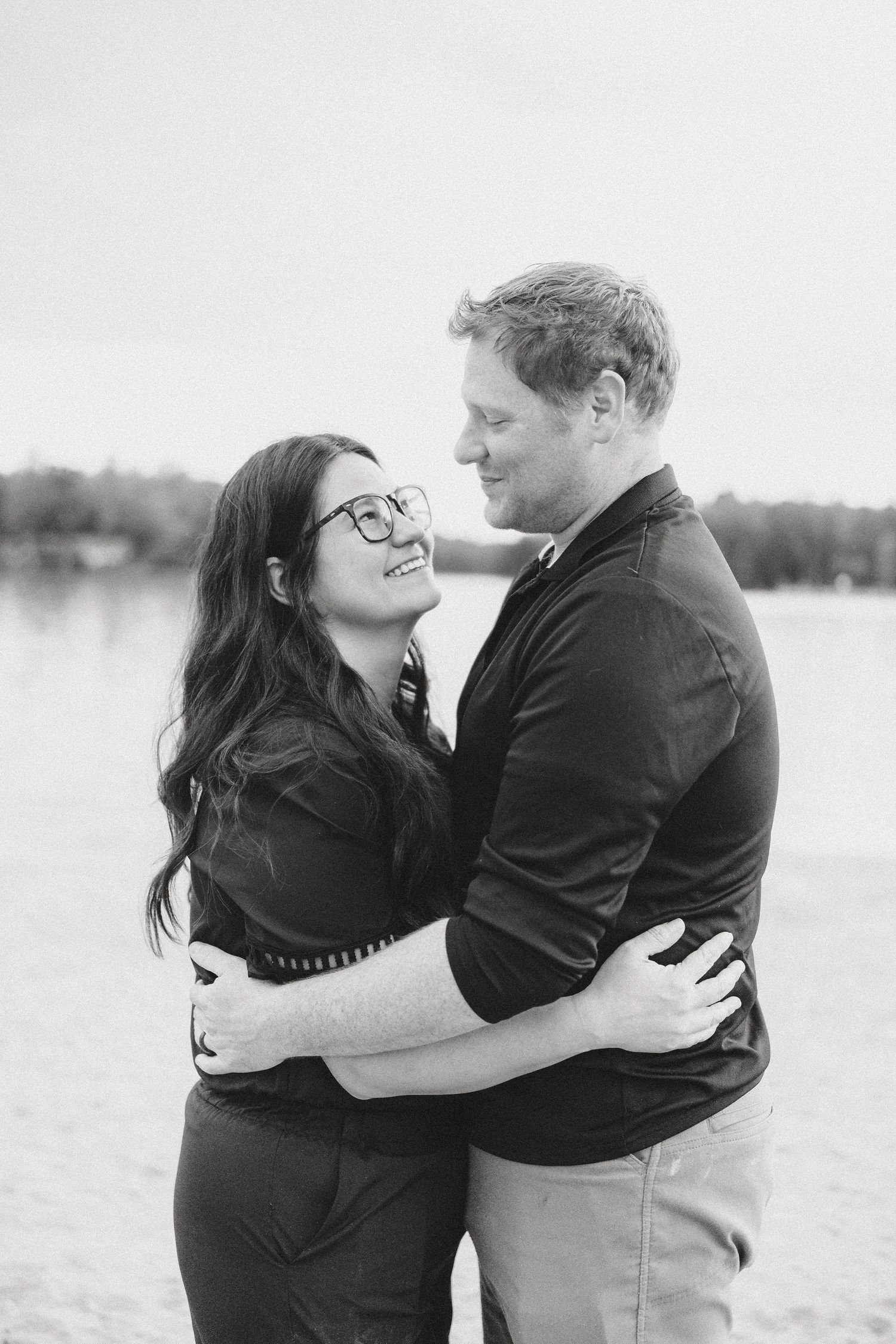 Black and white photo of a smiling couple embracing on a beach, woman in glasses looking up at man near a lake.
