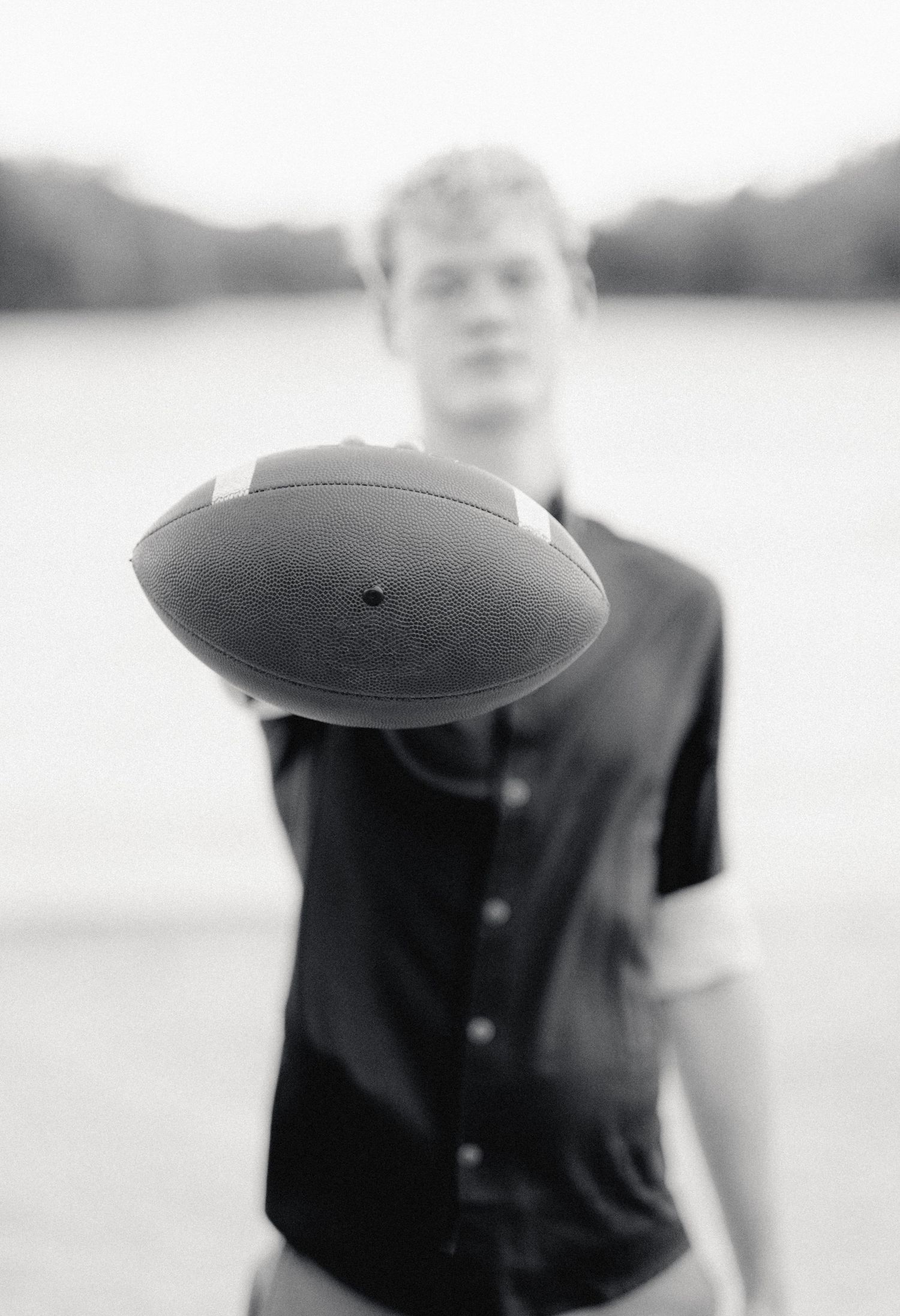 Black and white photo of a young man holding a football toward the camera outdoors near a lake.