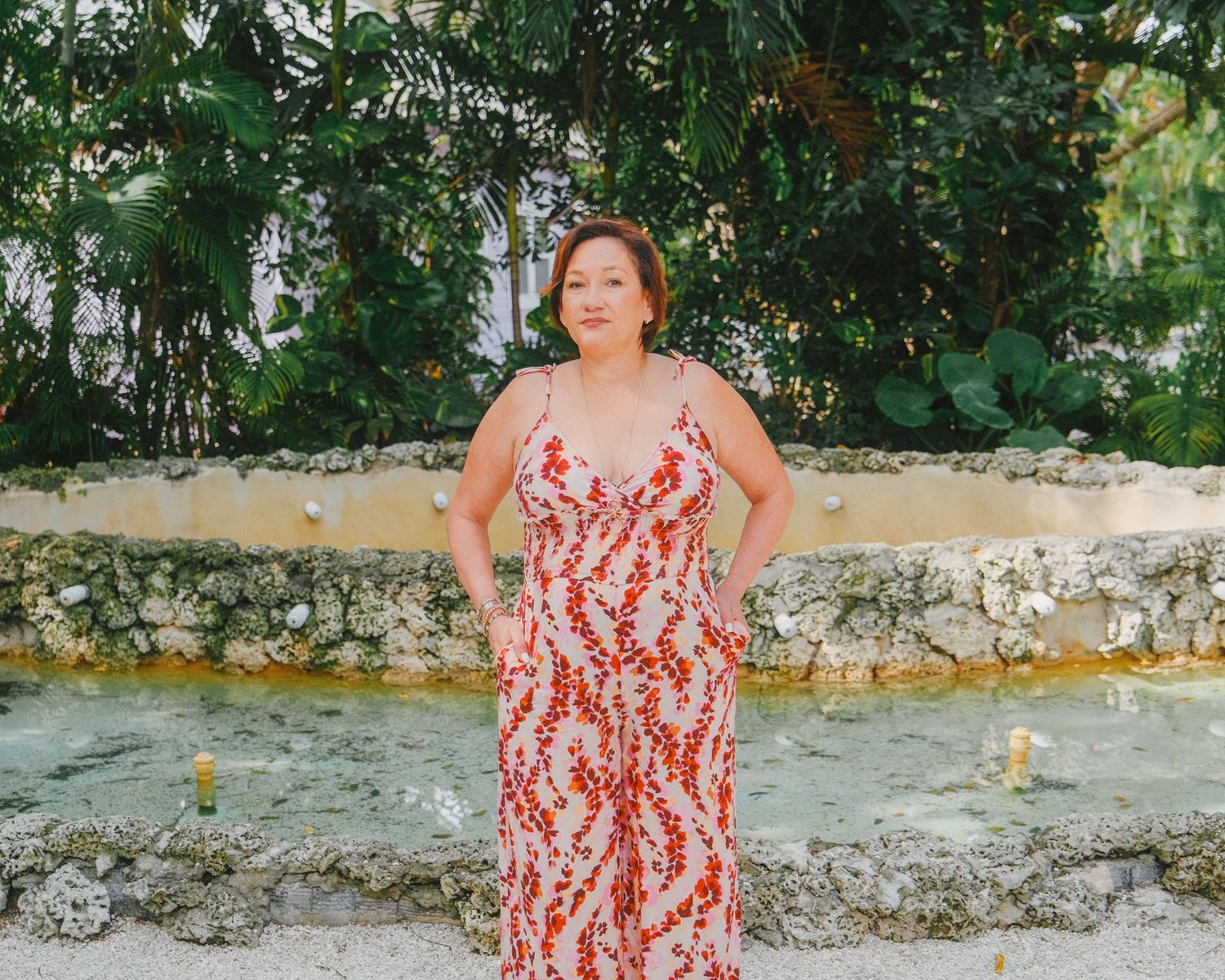 A person in a red and white floral maxi dress stands confidently against a stone wall with lush greenery in the background.