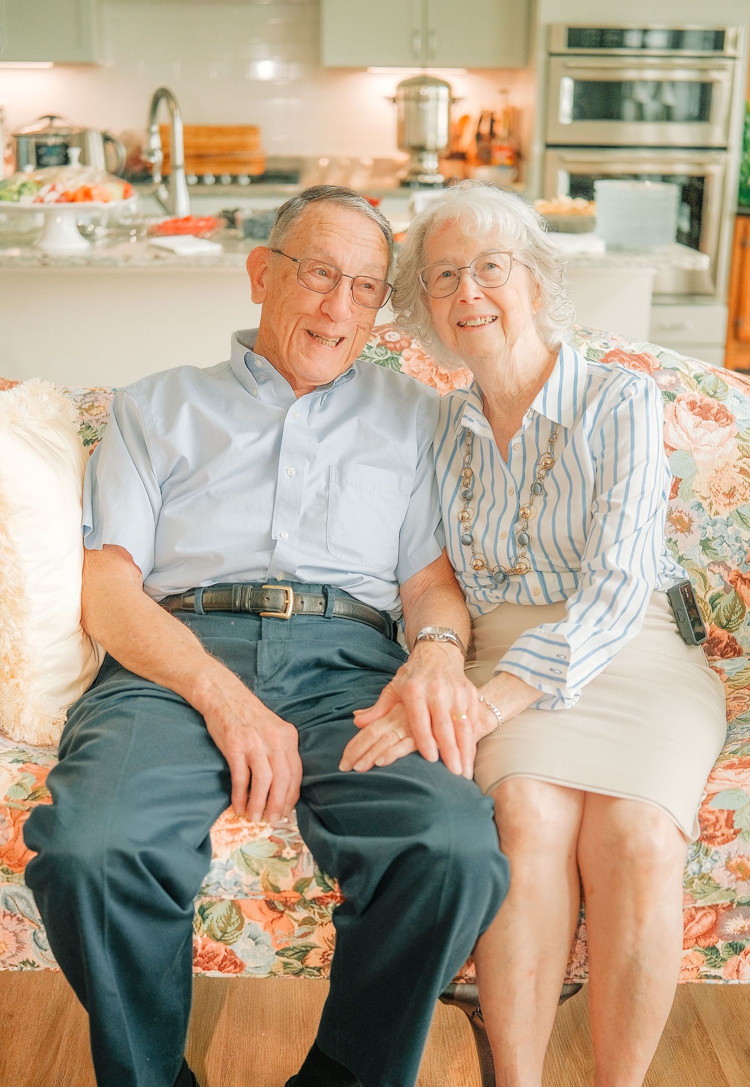 Elderly couple smiling and holding hands on a floral sofa in a bright modern kitchen background.