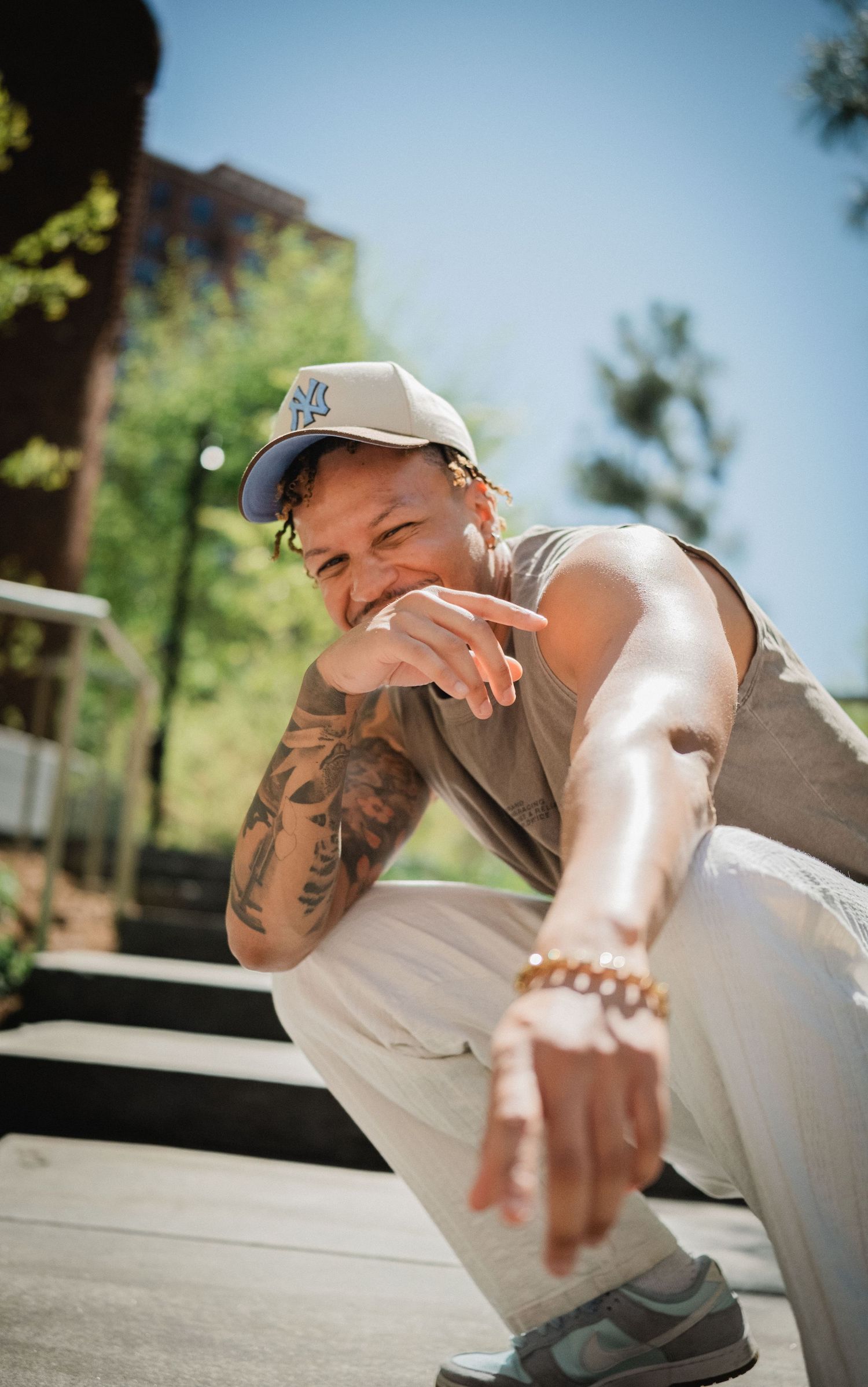 Tattooed man in NY Yankees cap crouching outdoors, smiling, wearing gold bracelet and gray sneakers on sunny day.