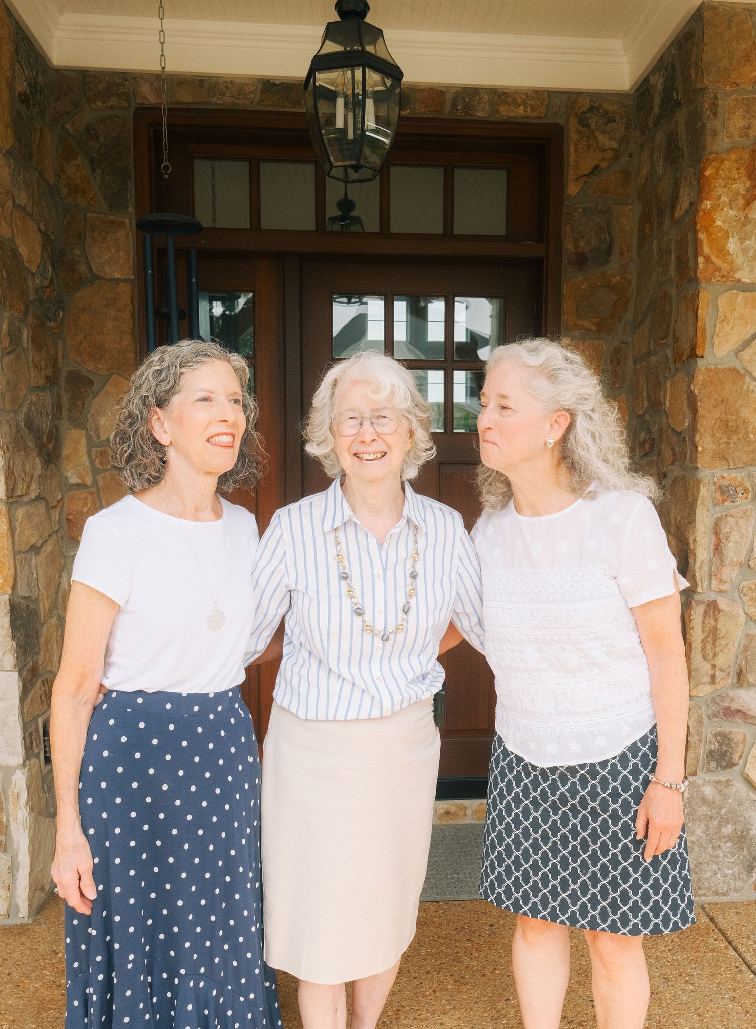 Three smiling older women with gray hair pose together outside a stone home entrance wearing casual summer outfits.