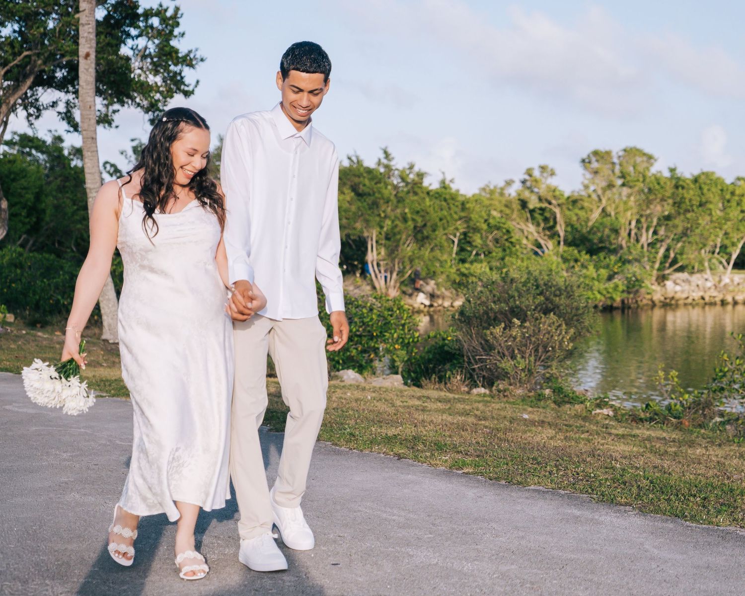 A couple in all white outfits holds hands while walking along a lakeside path during a romantic photoshoot.