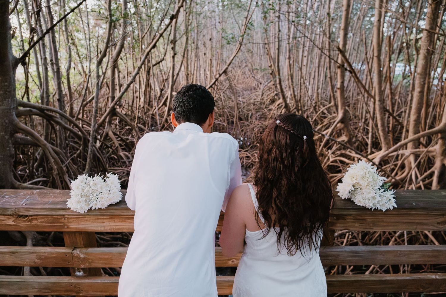 A couple in wedding attire sits together on a wooden bench in a rustic outdoor setting during autumn.
