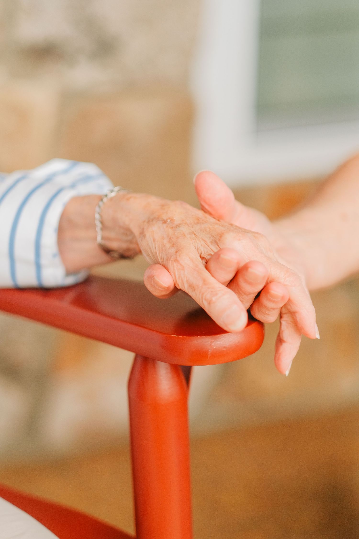 An elderly person's wrinkled hand resting on a red chair rail, gently held by a younger caregiver's smooth hand.