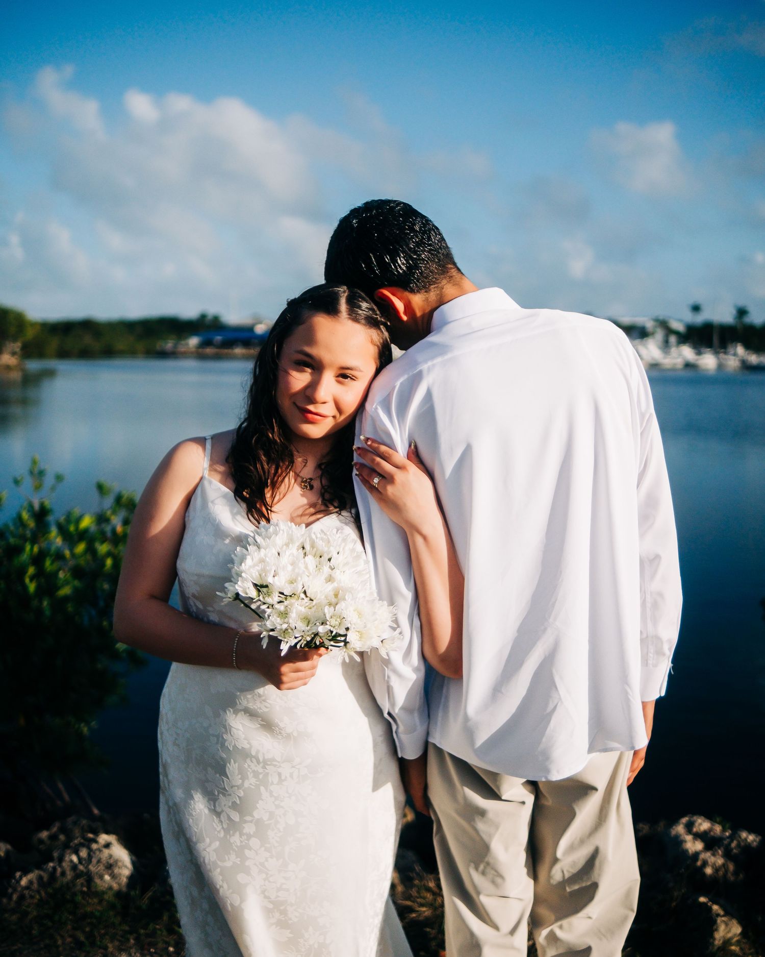 Bride in white lace gown holding bouquet stands with groom in white shirt by waterfront on sunny wedding day.