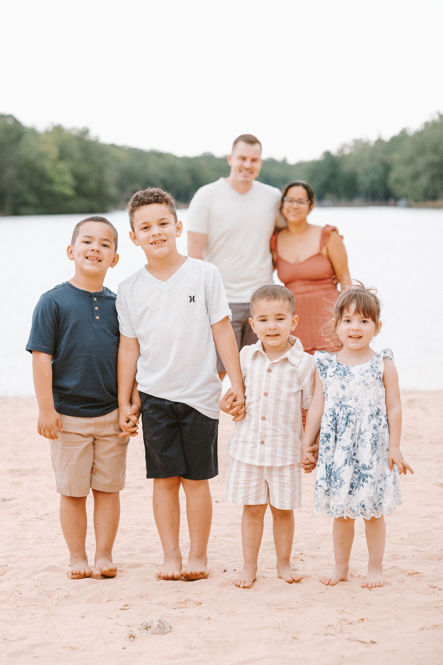 Family of six poses on sandy beach, two parents stand behind four young children holding hands near a serene lake.
