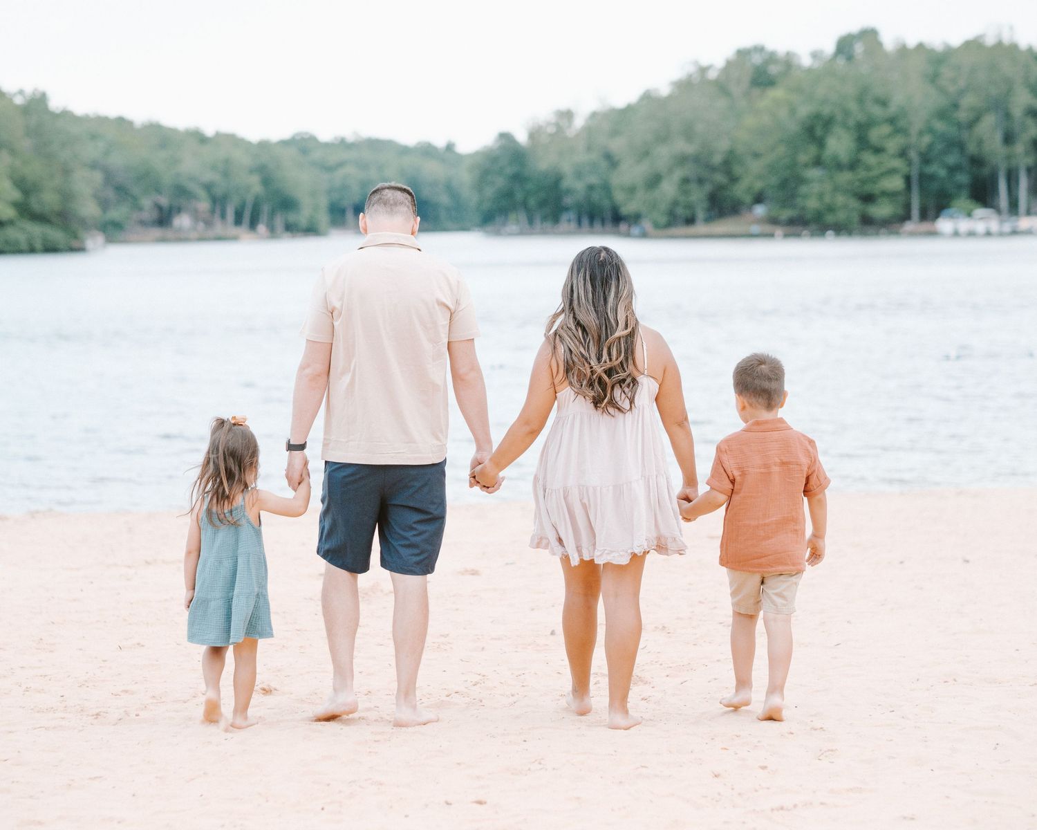 Family of four holding hands on sandy beach shore with lake and trees in background.