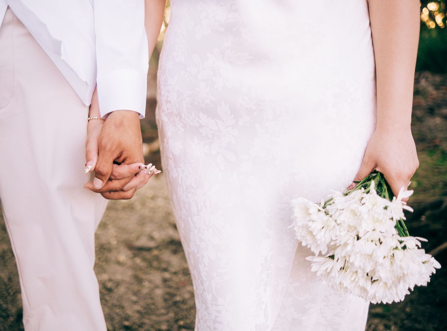 Close-up view of a bride in white lace gown holding white flowers while holding hands with someone in white attire.