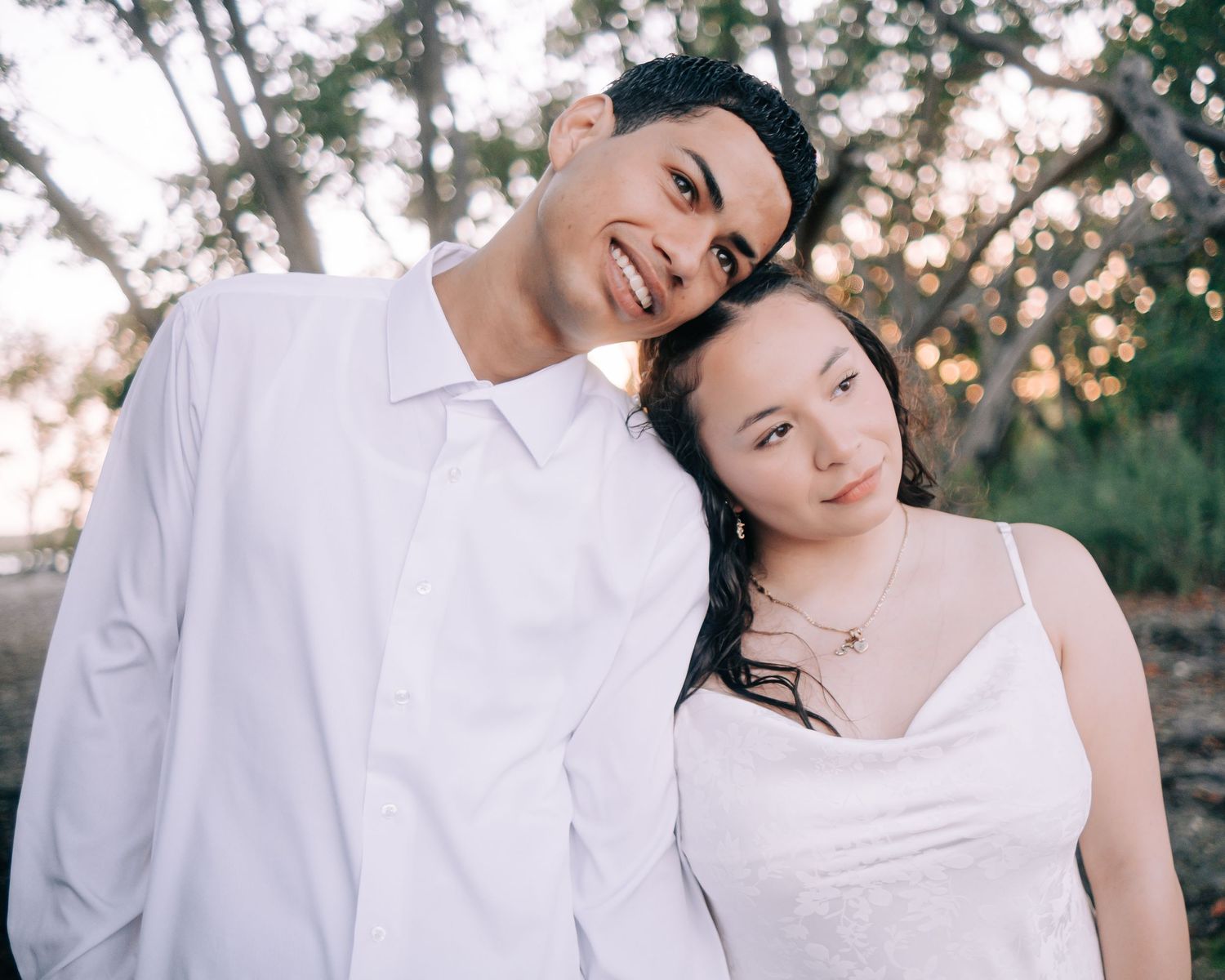 A couple in white attire smiling together outdoors during golden hour with trees in the background.