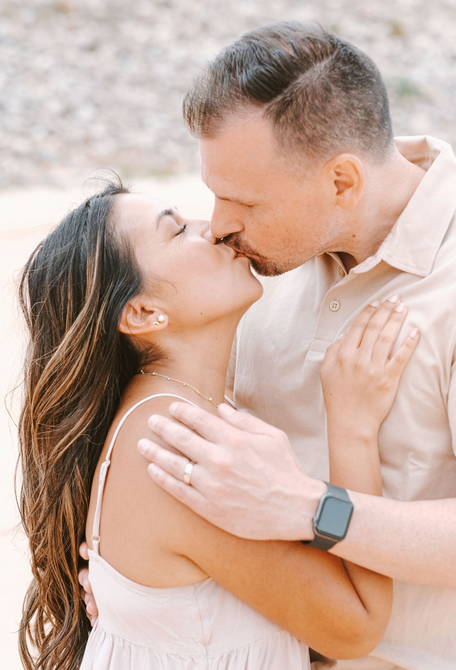 A couple shares a tender kiss outdoors, the woman wearing a pink dress and engagement ring.