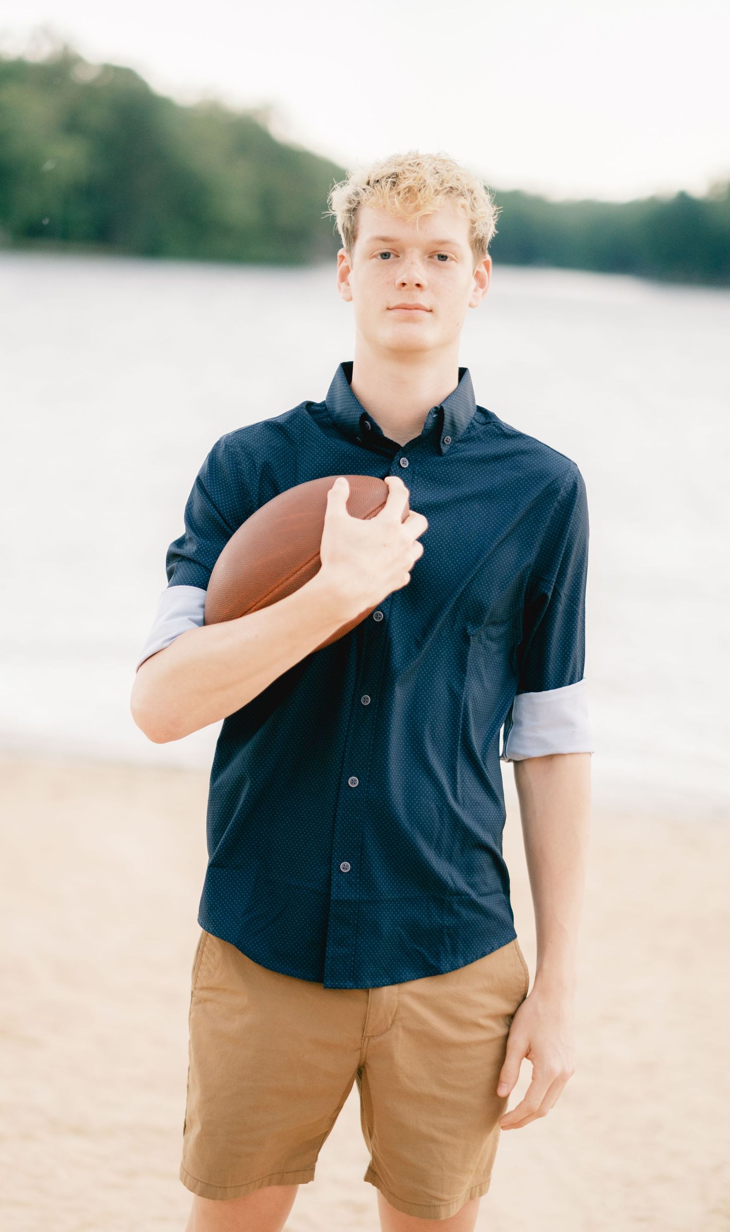 Young man in navy shirt holding a football on a sandy beach with water and trees in the background.