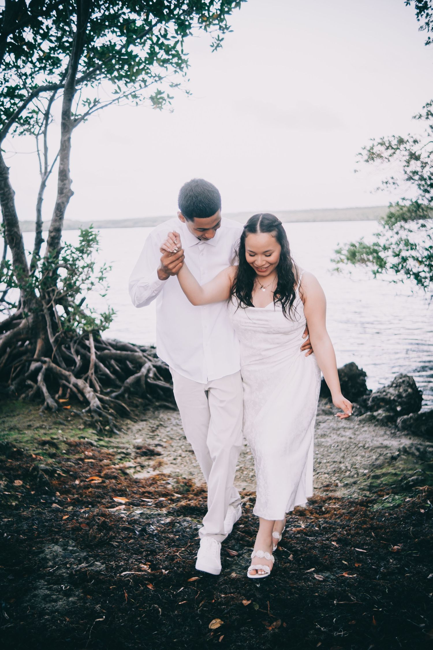 A couple in white attire walking and laughing together along a scenic waterfront path with mangrove trees.