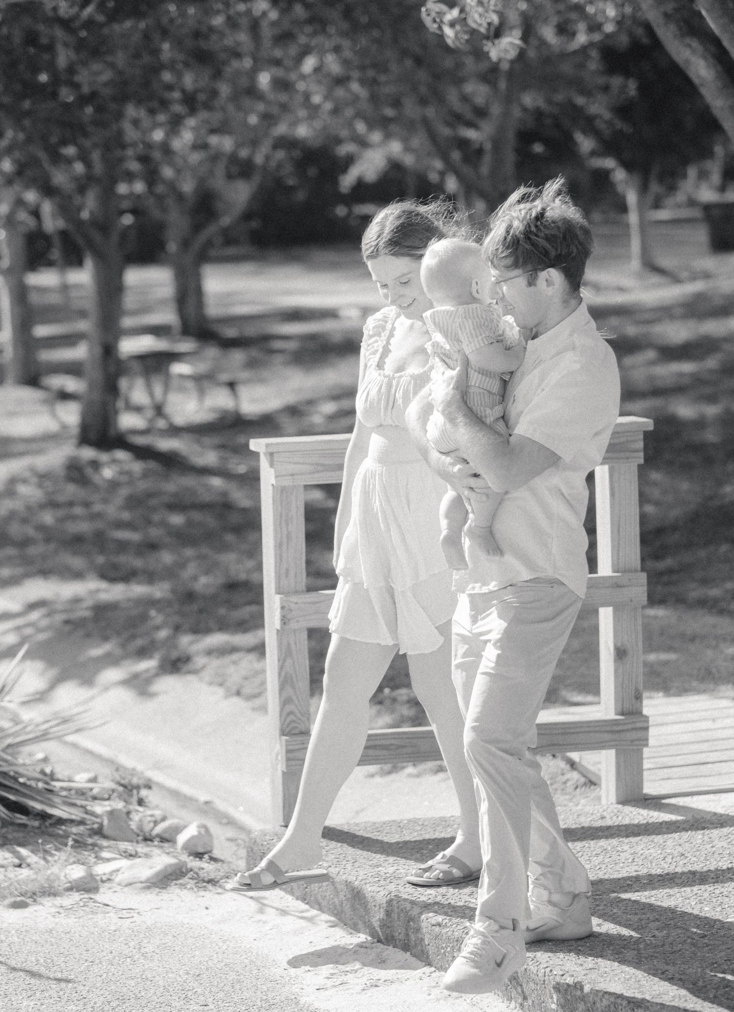 Black and white photo of a young family with parents holding a baby while walking through a sunny park.