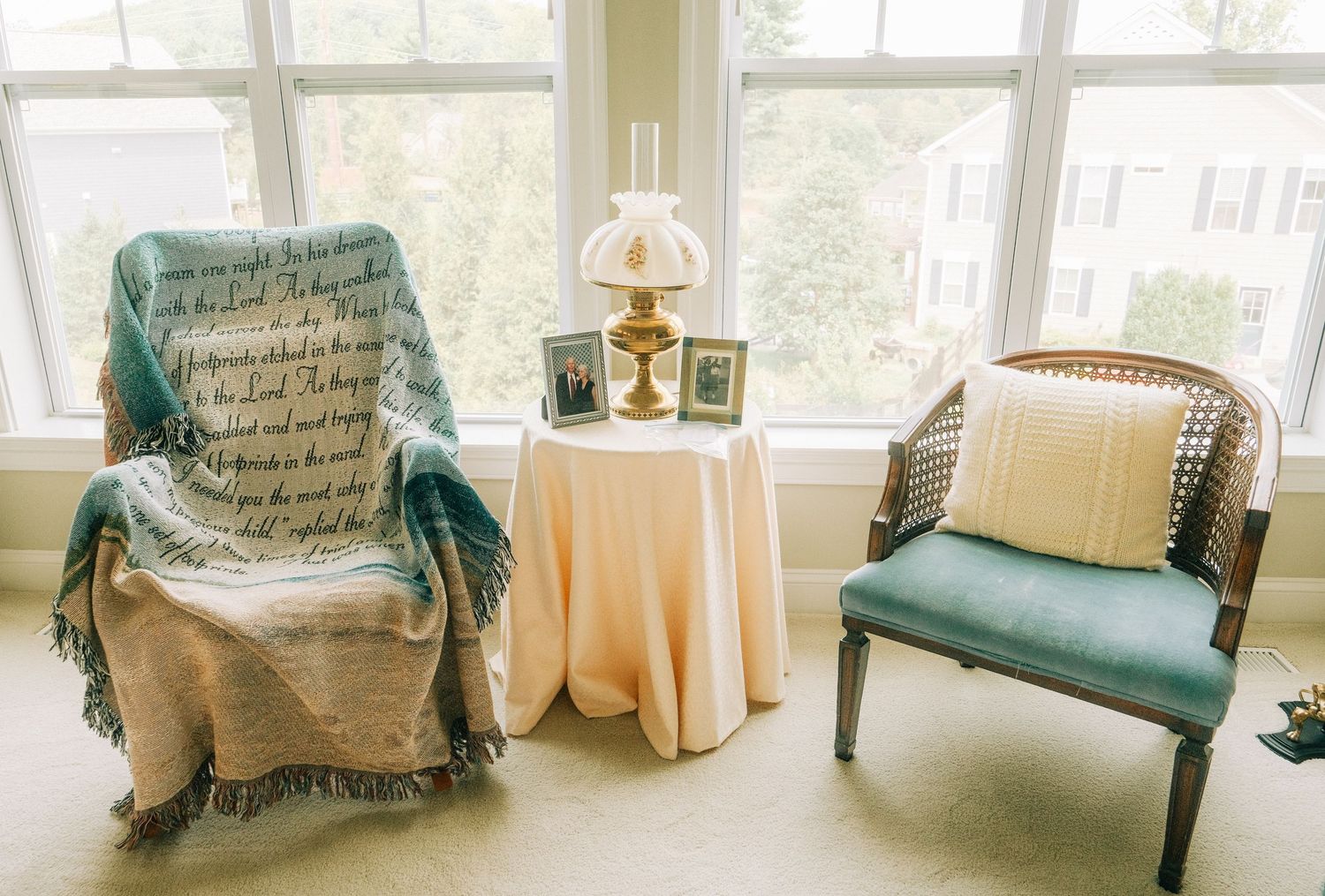 Cozy sitting area with two chairs, a draped throw blanket, lamp, and framed photos on a round table by bright windows.