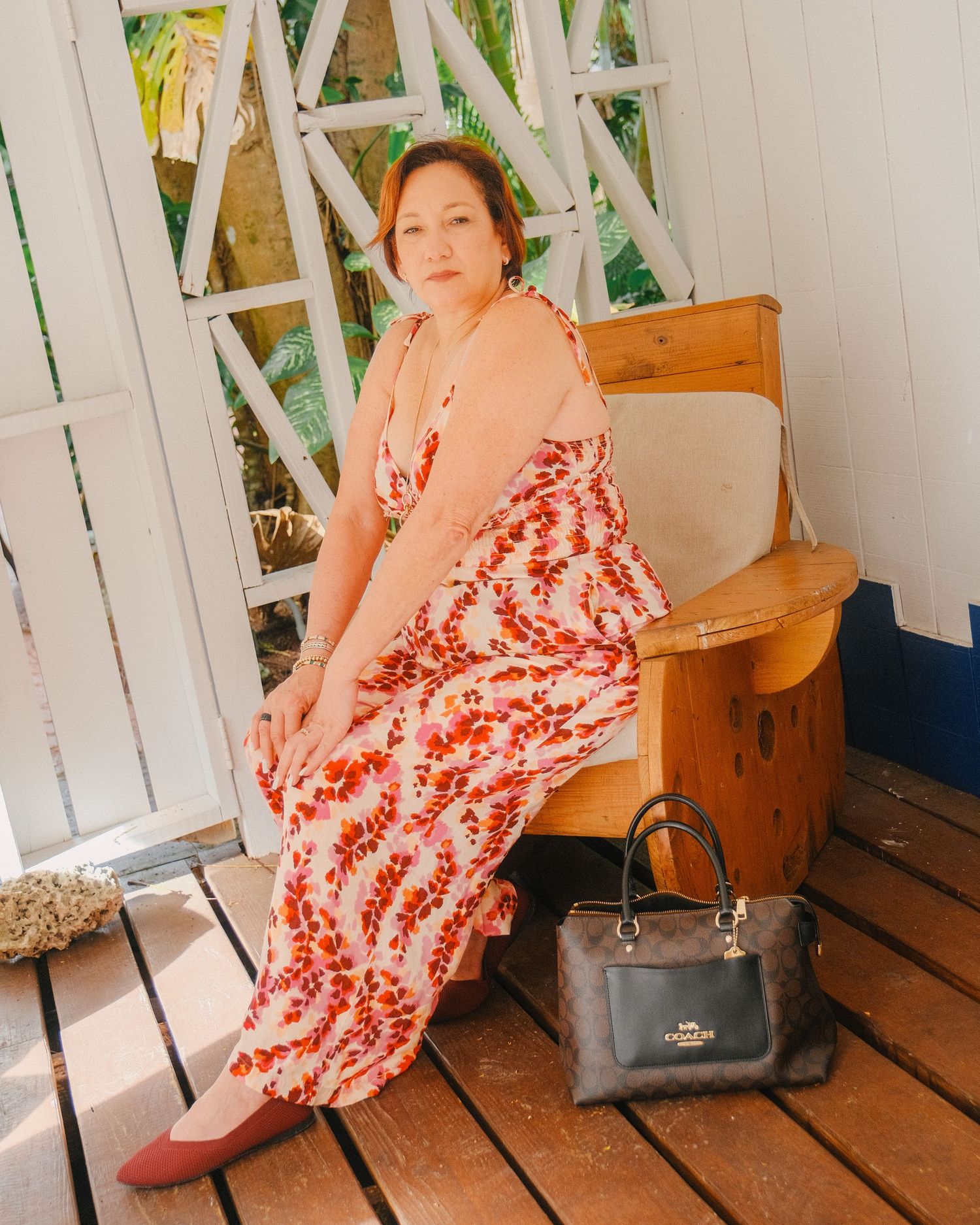 Someone wearing a peach cardigan and red floral maxi dress sits on a wooden porch with a gray handbag nearby.