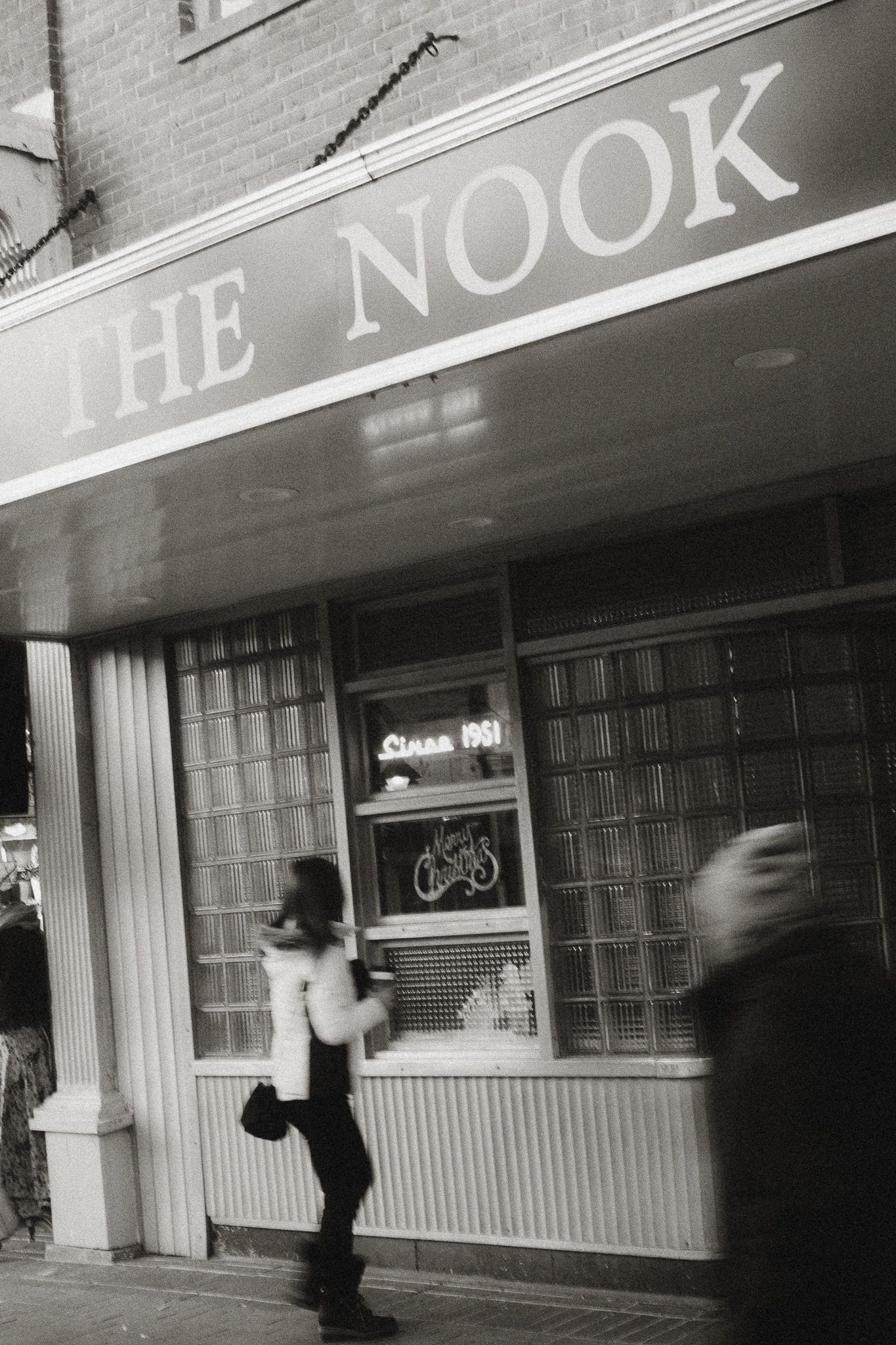 Black and white photo of The Nook storefront with blurred pedestrians walking past.