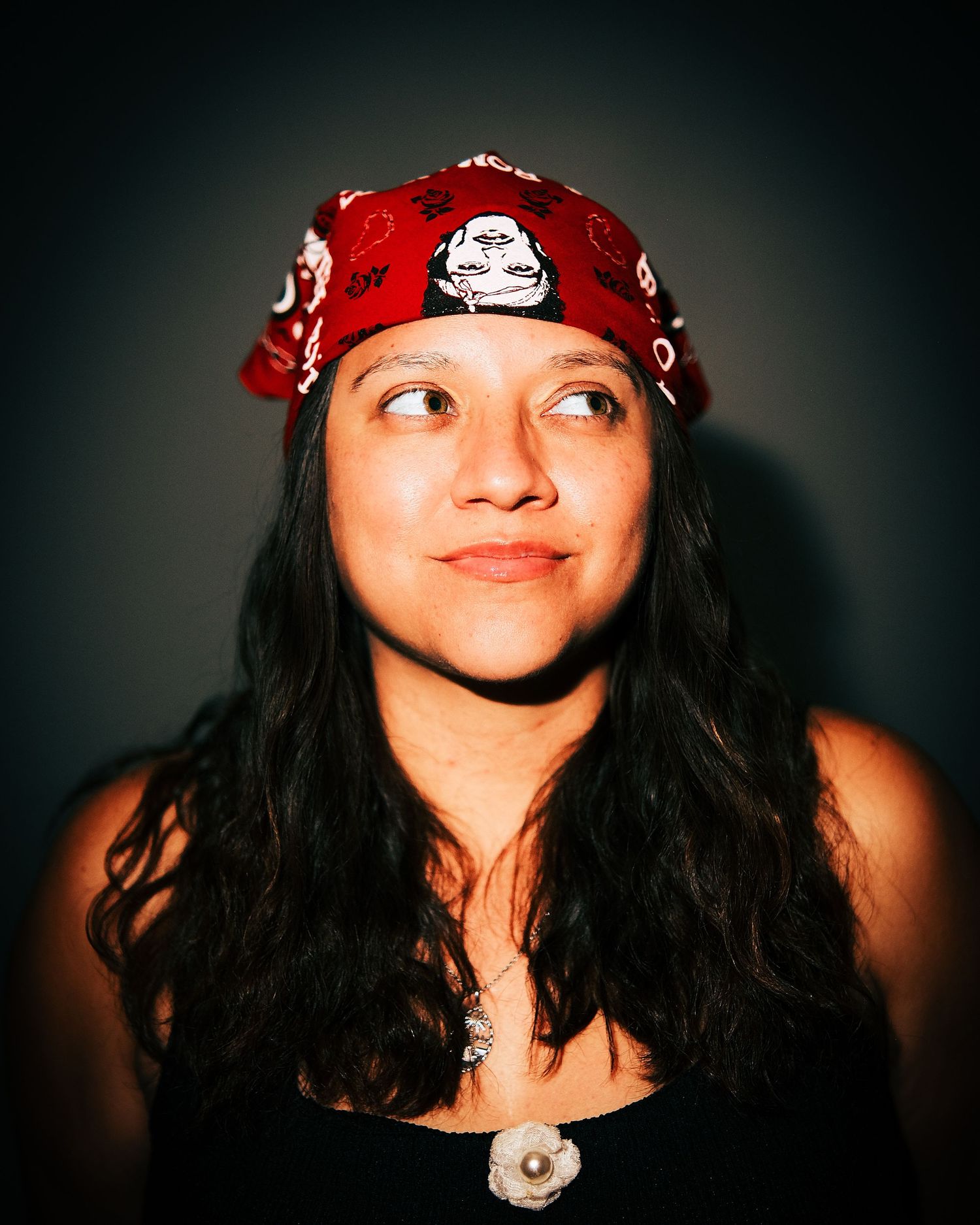Young woman wearing a red bandana headband with long dark hair, looking upward thoughtfully against a dark background.