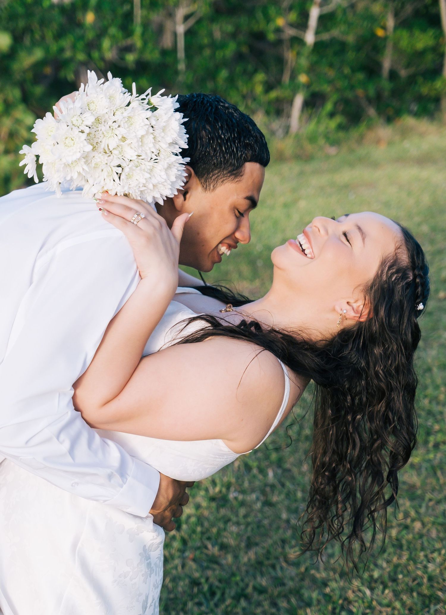 A romantic couple in white embraces and laughs joyfully on grass with white flowers.