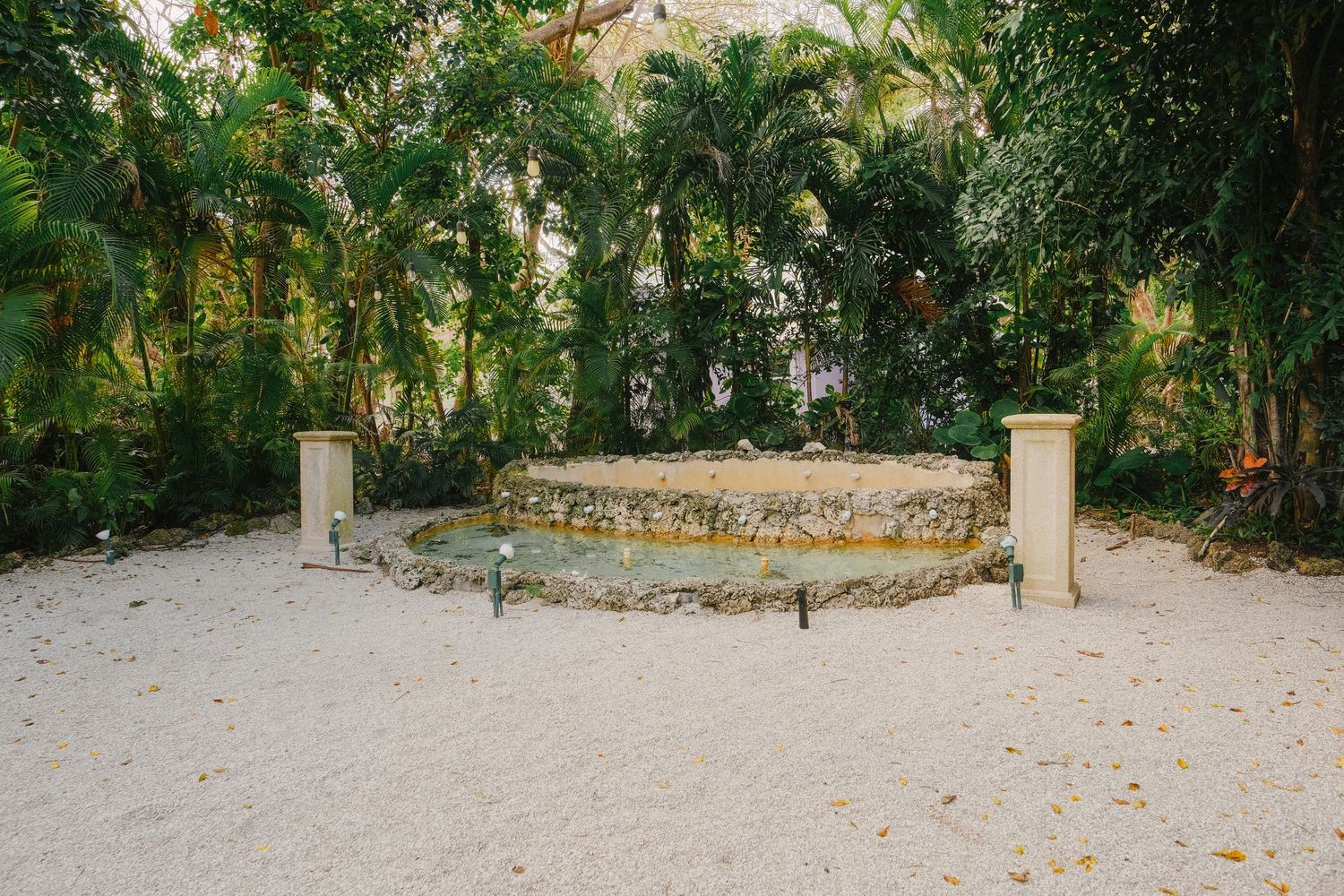 Stone pillars mark the entrance to a winding garden path surrounded by lush tropical foliage and palm trees.