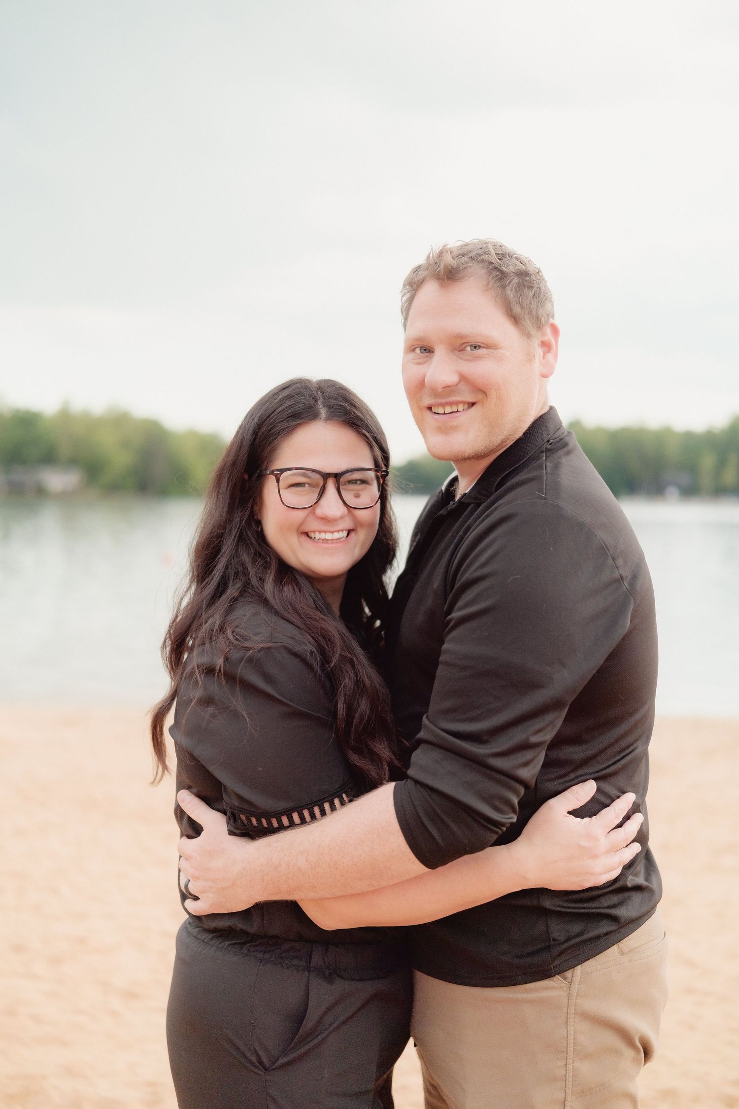 A smiling couple embraces on a sandy beach, both wearing black outfits, with a lake and trees in the background.