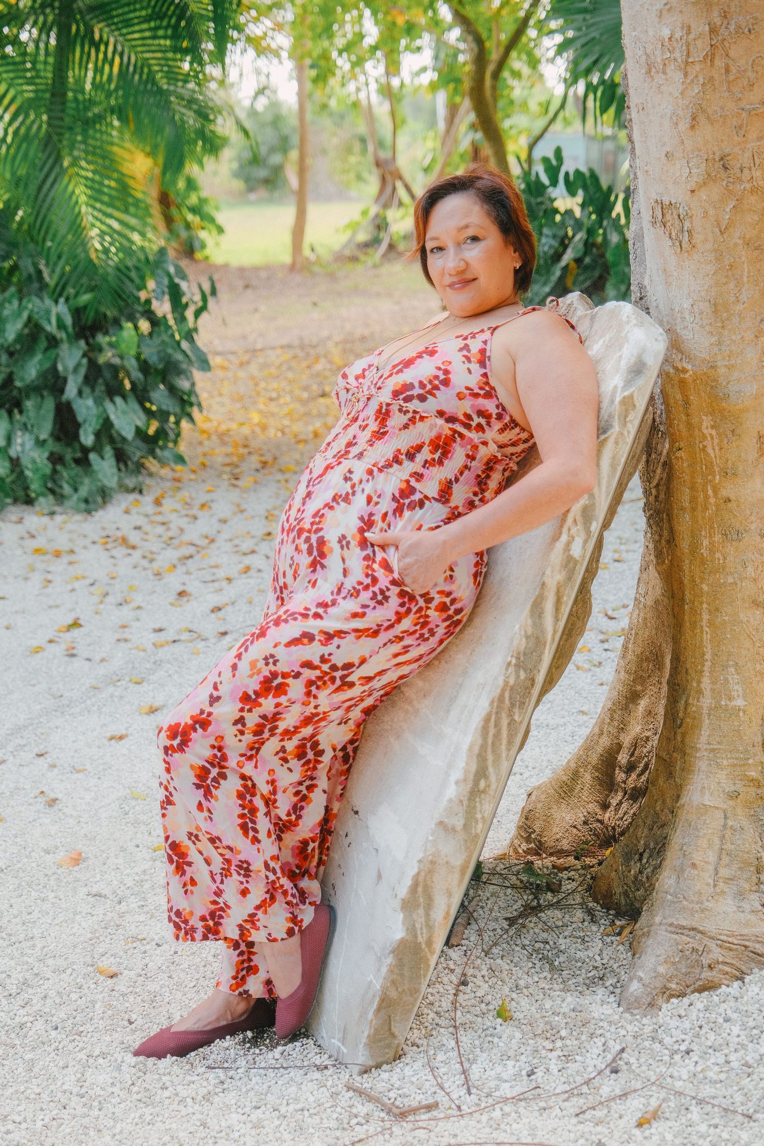 A child in a red floral dress sits against a tree trunk in a sunny outdoor setting with fallen leaves on the ground.