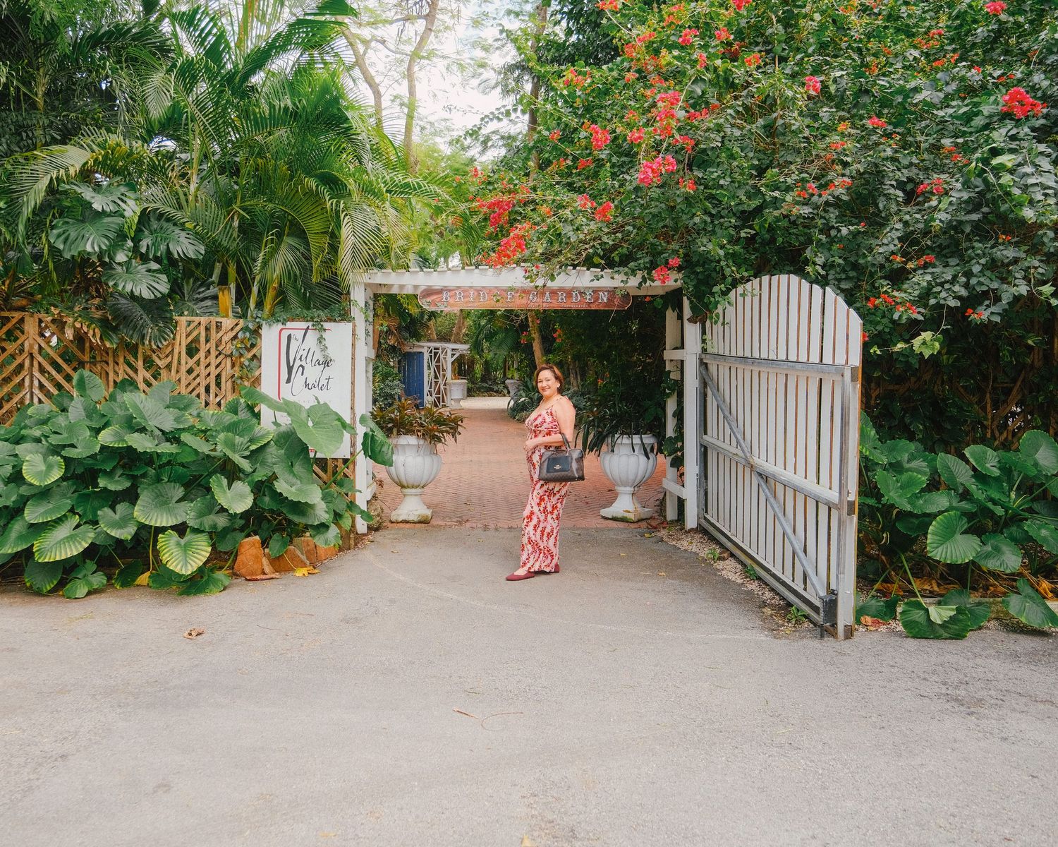 A garden pathway lined with white picket fences and vibrant red flowering plants leads to a tropical paradise.