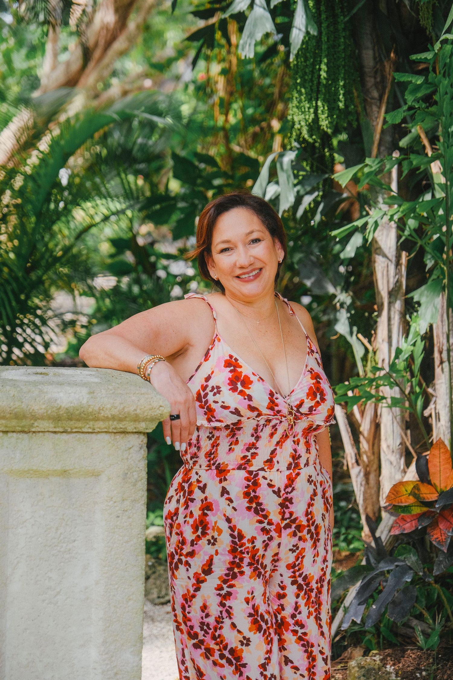 A person in a red and white floral dress poses outdoors against a stone column surrounded by lush greenery.