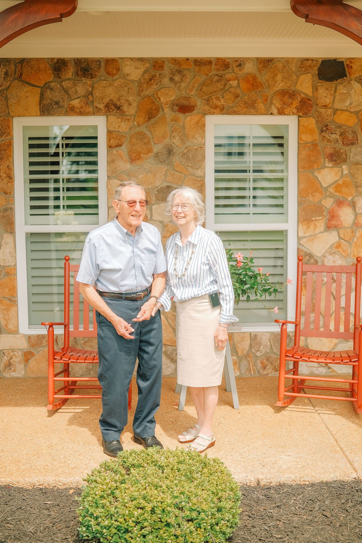 Elderly couple smiling together on a stone house porch with red rocking chairs on a sunny day.