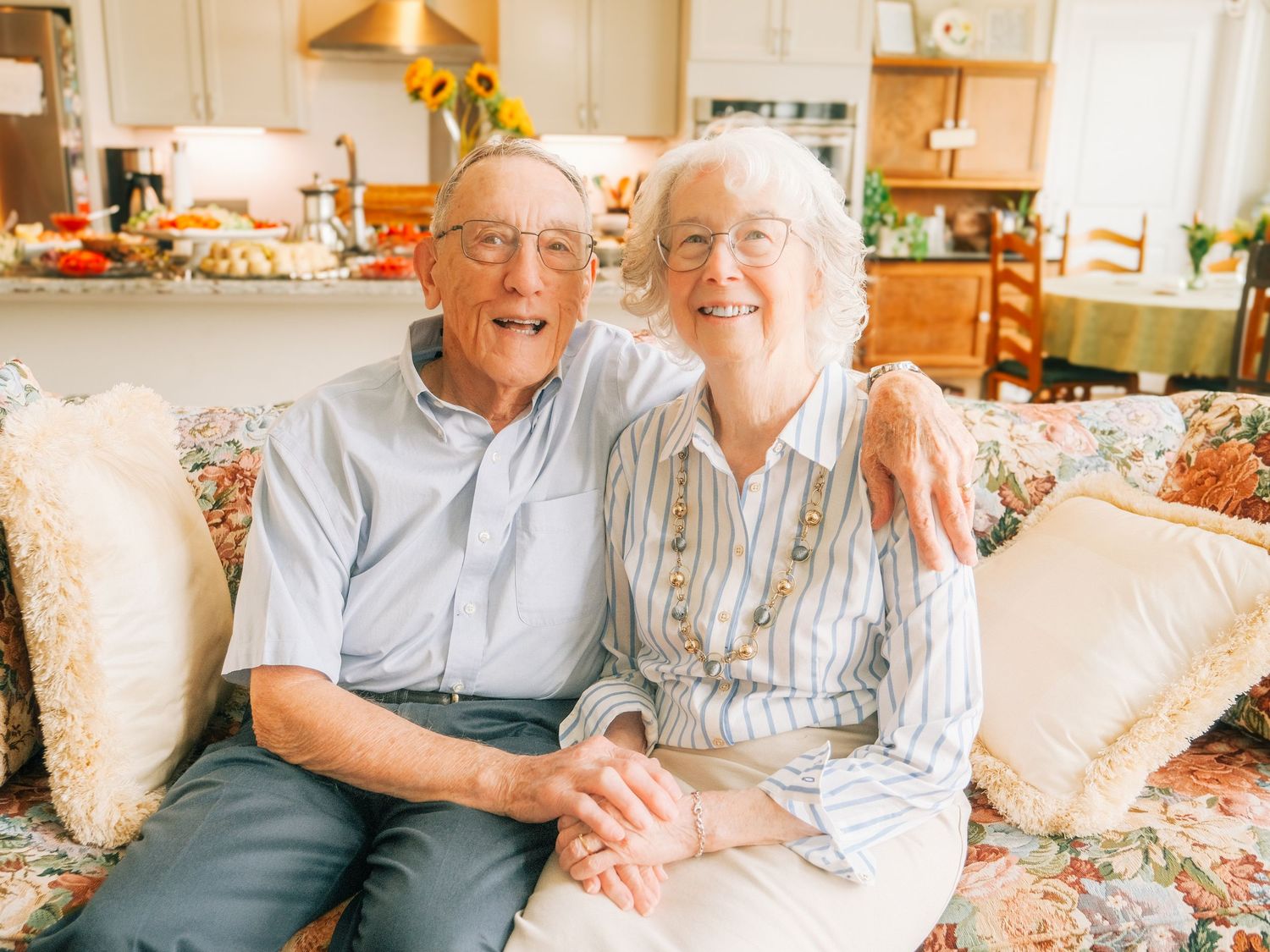 Happy elderly couple sitting together on a floral sofa, smiling warmly in a bright home setting.