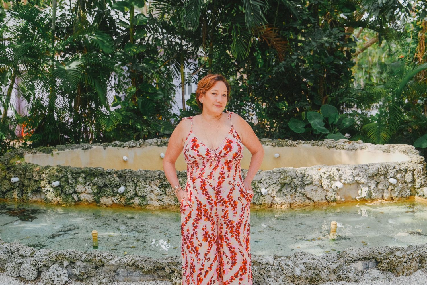Someone in a red and white floral sundress poses on a stone wall in a lush outdoor garden setting.