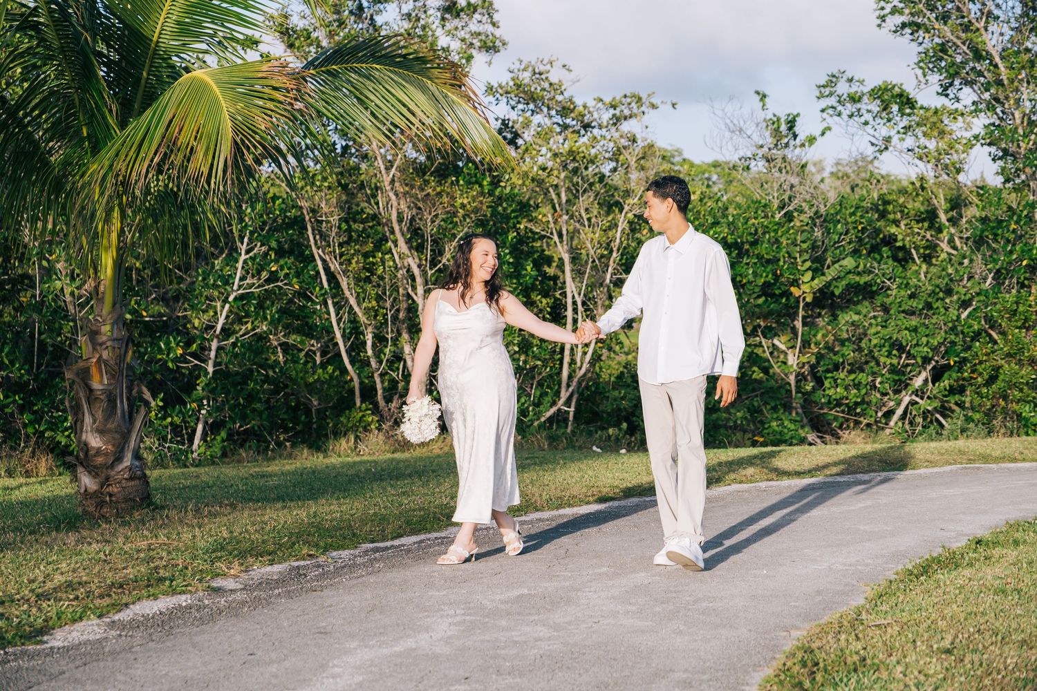 A couple in matching white outfits walks together on a path surrounded by tropical palm trees and greenery.