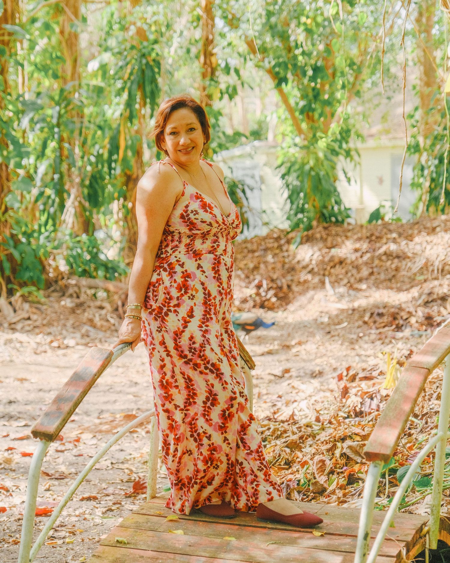 A person in a red and white floral dress sits on a wooden bench in a lush garden setting.