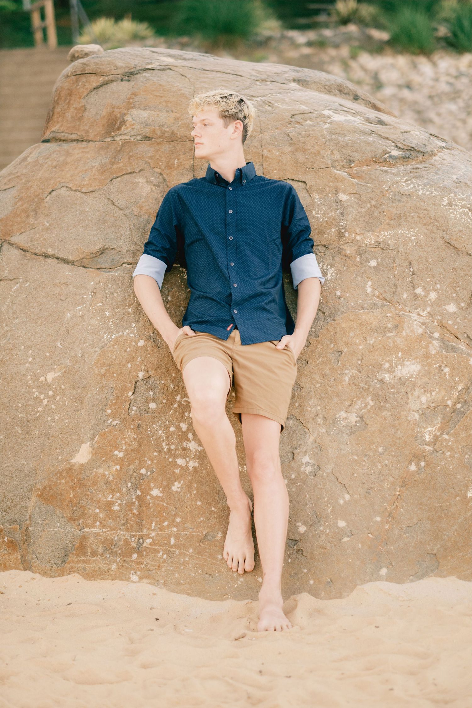 Young man in navy shirt and tan shorts leaning against a large rock on a sandy beach.