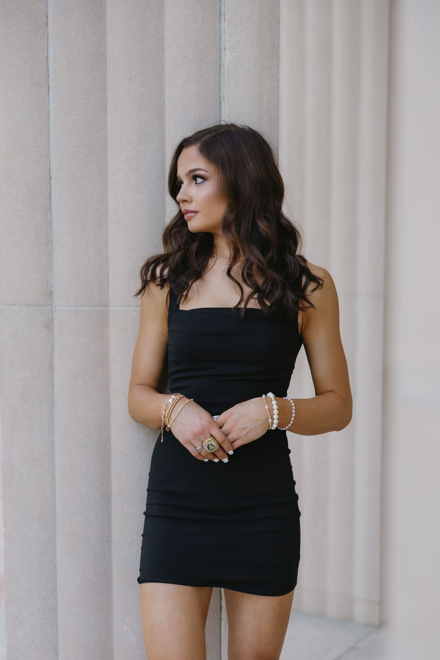Figure in black dress poses elegantly against white architectural columns in outdoor photoshoot.