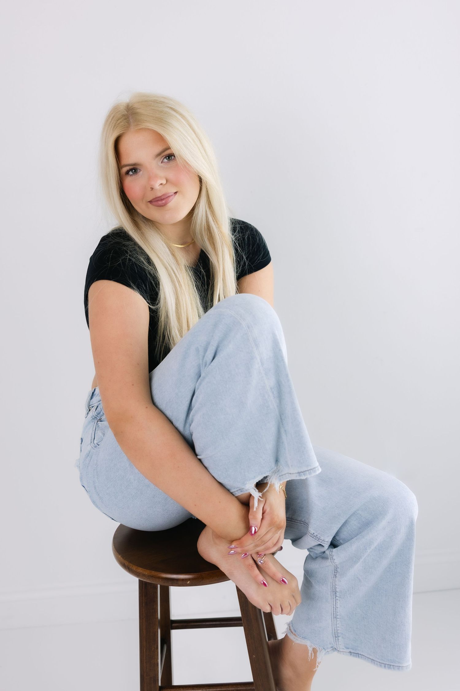 Model in light denim and black top poses casually on wooden stool in bright studio setting.