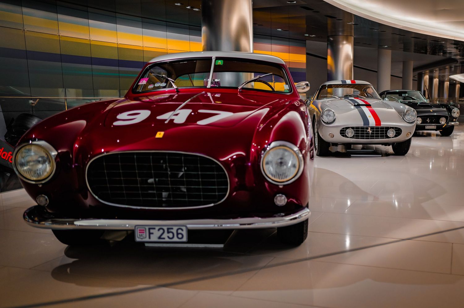 Vintage Ferrari race cars on display in a museum, featuring a red Ferrari numbered 947 with license plate F256.